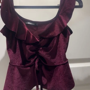 Vinröd glittrig topp från Gina Tricot - Säljer en vinröd glittrig topp från Gina Tricot med volang vid axlarna och snörning framtill. Toppen är ärmlös och har en croppad passform som ger en snygg siluett. Perfekt för att sticka ut med lite extra glam.