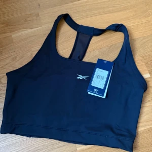 Sporttopp från Reebok - Sportig svart sporttopp från Reebok med brottarrygg och meshdetalj baktill. Toppen har en stilren design med Reebok-logga framtill och är tillverkad i ett stretchigt syntetmaterial som passar perfekt för träning.