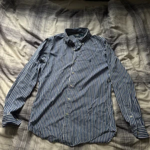 Randig skjorta från Ralph Lauren - Säljer en blåvit randig skjorta från Polo Ralph Lauren i custom fit. Skjortan har klassisk krage, långa ärmar och knäppning framtill. Ikoniska broderade loggan på bröstet. Perfekt för dig som gillar stilrena och tidlösa plagg.