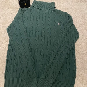 Grön kabelstickad polotröja från GANT - Mörkgrön kabelstickad polotröja från GANT i premium bomull. Tröjan har hög krage, lång ärm och broderad GANT-logga på bröstet. Klassisk och stilren modell med ribbade muddar och strukturstickat mönster.