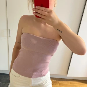 Ljusrosa ribbad bandeau tubtopp - Säljer en ljusrosa bandeau tubtopp med ribbad struktur. Toppen är axelbandslös och sitter tight mot kroppen, perfekt för varma dagar. 