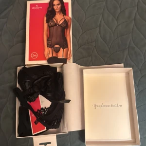 Svart spetskorsett från Obsessive - Säljer en svart korsett från Obsessive med transparent mesh, spetsdetaljer och snörning i ryggen. Smala axelband och dekorativa spetskanter. Medföljer matchande stringtrosa. Perfekt för dig som vill ha något extra snyggt och vågat.