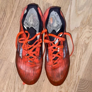 Adidas F50 - Säljer ett par röda Adidas fotbollsskor med svarta och silvriga detaljer. Skorna har snörning och platt sula med fasta dobbar för bra grepp på planen. Ovandelen är i syntetmaterial och designen är sportig och modern.