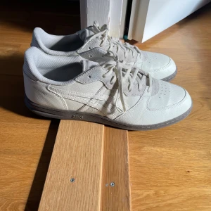 Vita Asics Skyhand sneakers - Säljer ett par vita Asics Skyhand sneakers med klassisk design. Skorna har snörning, rund tå och platt sula. Ovandelen är i skinn och textil, med Asics logga på sidan och grå detaljer. Perfekta för dig som gillar retrostil och bekväma sneakers.