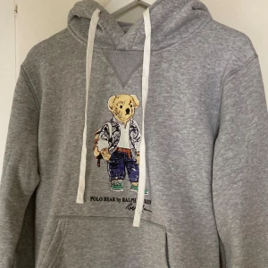 Grå hoodie Polo Bear Ralph Lauren - Grå hoodie från Polo Ralph Lauren med ikoniskt Polo Bear-tryck på bröstet. Tröjan har huva med vita snören och en stor magficka. Mjuk och skön bomullskänsla,  storlek M                                                använd en gång, 2 h max, aldrig tvättad ca 2 månader gammal