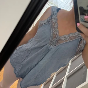 Ljusblå linnetopp med spetsdetaljer - Supersöt ljusblå linnetopp med breda axelband och v-ringning. Toppen har fina spetsdetaljer med blommönster längs ringningen och axelbanden. Luftig och flowy passform, perfekt för varma dagar.