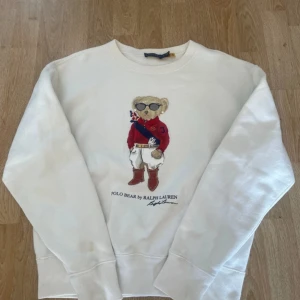 Vit sweatshirt Polo Bear Ralph Lauren - Vit sweatshirt från Ralph Lauren med ikoniska Polo Bear-trycket framtill. Tröjan har rund hals, långa ärmar och ribbade muddar. Klassisk och stilren design med broderad nalle i rött och blått. Perfekt för dig som gillar preppy stil. Har några små fläckar som jag har visat på bilden