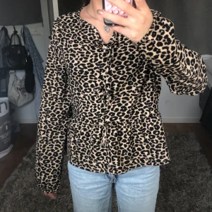 Leopardmönstrad blus från Kappahl - Säljer en snygg leopardmönstrad blus från Kappahl i beige och svart. Blusen har långa ärmar och flera knytningar framtill som cool detalj. Materialet är mjukt och lätt, perfekt för att lyfta vilken outfit som helst.