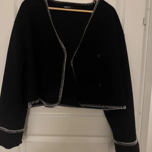 Svart cardigan jacka från Gina Tricot - Svart cardigan jacka från Gina Tricot med kontrasterande vita sömmar längs kanterna och ärmsluten. Jackan är croppad, har lös passform och knäpps med två knappar framtill. Perfekt för dig som gillar en stilren men ändå unik look.