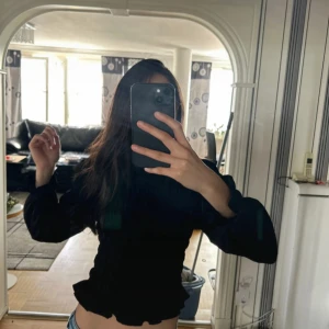 Svart smockad offshoulder blus - Säljer en svart blus med smockad kropp och långa puffärmar. Toppen har offshoulder-modell och volangkant nertill för en extra söt touch. Perfekt att styla med jeans eller kjol för en trendig look.