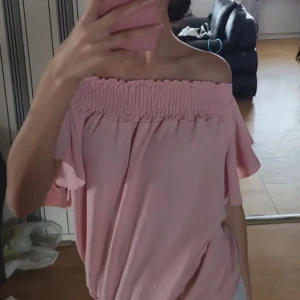 Ljusrosa offshoulder blus - Säljer en söt ljusrosa offshoulder blus med smockad överdel och vida, korta ärmar. Toppen har en luftig och flowy passform, perfekt för varma dagar. Superfin att styla med jeans eller kjol för en trendig look.