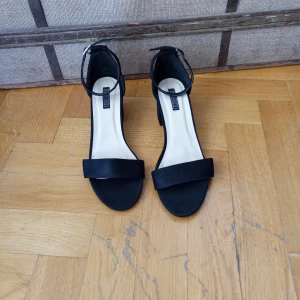 Svarta sandaler med klack från Eko Shoes - Stilrena svarta sandaler från Eko Shoes med bred rem över tårna och justerbart ankelband med spänne. Skorna har en stabil blockklack och öppen tå, perfekta för att lyfta din outfit. Materialet är mockaliknande och ger en lyxig känsla.