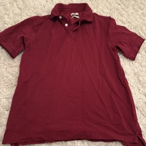 Vinröd pikétröja från Gant, stl S - Säljer en vinröd pikétröja från Gant i 100% bomull. Klassisk modell med krage, två vita knappar och diskret broderad logga på bröstet. Perfekt för en clean och stilren look. Tröjan har korta ärmar och är riktigt skön att bära.