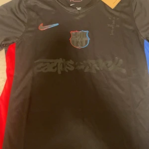 FC Barcelona x Cactus Travks Scott, Jack Nike tröja - Säljer en svart FC Barcelona x Cactus Travis Scott, Jack fotbollströja från Nike. Tröjan har korta ärmar, klubbmärke med blå och röd kant, samt Cactus Jack-tryck på bröstet. Sidorna har röda och blå paneler. Tillverkad i lätt och ventilerande material.