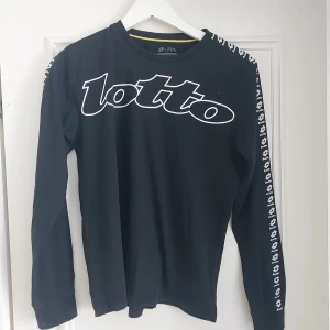 Svart långärmad tröja från Lotto,storlek 13-14år - Svart långärmad tröja från Lotto med stort vitt logotryck över bröstet och vita logodetaljer längs ärmarna. Tröjan har rund halsringning och är tillverkad i mjuk bomull, perfekt för en sportig och avslappnad stil.