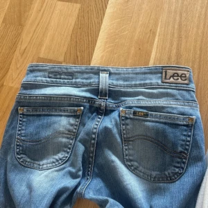 Blå bootcut jeans från Lee - Snygga blå jeans från Lee med bootcut-modell och klassisk femficksdesign. Jeansen har låg midja, ljusa slitningar framtill och Lee-logga på bakfickan. Perfekta för dig som gillar en avslappnad och trendig look. De är nästan helt nya!