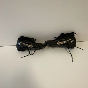 Svarta Nike fotbollsskor med guldiga detaljer - Säljer ett par svarta Nike fotbollsskor med snygga guldiga och vita detaljer. Nästan helt oanvända har tränat 5 träningar med dom pga att jag tröttnade på fotboll och sen har dom stått hemma i 2 år. 