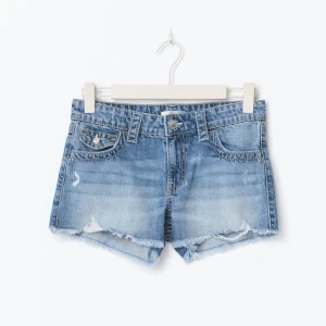 Blå jeansshorts med fransig kant - Säljer ett par snygga blå jeansshorts med slitna detaljer och fransig kant nertill. Shortsen har klassisk femficksdesign, knapp och dragkedja framtill samt en cool tvättad look. Perfekta för varma dagar och enkel att matcha med allt.