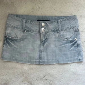 Ljusblå jeanskjol från Olala - Säljer en ljusblå jeanskjol från Olala med klassisk femficksdesign och snygga detaljer på bakfickorna. Kjolen har silverfärgade knappar framtill och är i en kort, lågmidjad modell som passar perfekt till sommaren.