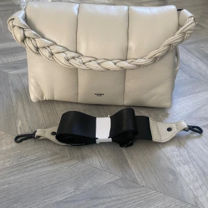 Beige quiltad axelväska från Ulrika - Ny snygg quiltad axelväska från Ulrika i färgen Kit,  skinnimitation med flätat handtag och avtagbar bred axelrem. Väskan har orange foder, dragkedja och flera innerfack för smart förvaring. Perfekt för dig som vill ha en trendig och praktisk väska.