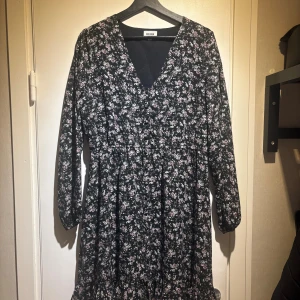Blommig svart klänning från Bubbleroom - Supersöt svart klänning från Bubbleroom med små blommor i vitt och lila. Klänningen har v-ringning, knappar framtill och långa ärmar. Den har en avslappnad passform och är perfekt för dig som gillar romantiska detaljer och blommigt mönster.