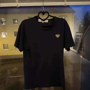 Svart Comme des Garçons Play t-shirt - Svart t-shirt från Comme des Garçons Play med klassiskt hjärta med ögon broderat i guld på bröstet. T-shirten har rund hals, rak passform och är tillverkad i mjuk bomull. Perfekt för dig som gillar stilrena och ikoniska plagg. Obs hål i tröjan kolla bild 3