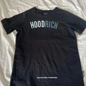 Svart t-shirt från Hoodrich - Svart t-shirt från Hoodrich med tryckt logga i vitt och blått framtill och texten 'FROM NOTHING TO SOMETHING' nedtill. Klassisk rund hals och korta ärmar. Skön och enkel stil, perfekt till jeans eller shorts.