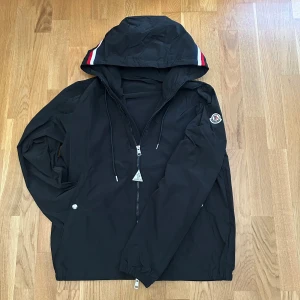 Svart vindjacka från Moncler - Säljer en svart vindjacka från Moncler med huva och dragkedja framtill. Jackan har Moncler-logga på ärmen och snygga röd-vita-blå ränder på huvan. Tillverkad i lätt polyester och har långa ärmar samt snörning i huvan. Perfekt för dig som vill ha en stilren och sportig look. Pris kan diskuteras||