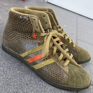 Adidas sneakers med ormskinnsmönster - Är i storlek 37, men passar mig som brukar ha 38-39! Unika Adidas sneakers i olivgrön mocka med ormskinnsmönstrade paneler och klassiska ränder i gult och orange. Högt skaft, snörning och röda Adidas-loggor på sidan och hälen. Perfekt för dig som vill sticka ut med coola detaljer och retrovibbar.