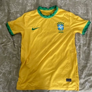Gul Brasilien fotbollströja! - Gul Brasilien fotbollströja från Nike med gröna detaljer på krage och ärmslut. Broderat CBF-märke och Nike-logga på bröstet. Tillverkad i lätt och ventilerande polyester, perfekt för match eller träning.