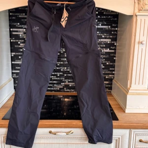 Svarta täckbyxor RevolutionRace S - Svarta täckbyxor från RevolutionRace i storlek S. Byxorna har dragkedjor vid knäna, snörning i midjan och praktiska fickor. Perfekta för utomhusaktiviteter och tillverkade i slitstarkt syntetmaterial.