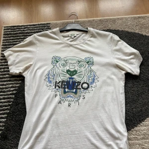 Kenzo t-shirt med tigertryck - Två snygga Kenzo t-shirts med ikoniskt tigertryck på bröstet. Ena är vit med grönt och blått tryck, andra är marinblå med orange och blått tryck. Båda har rund hals och korta ärmar, tillverkade i mjuk bomull. Perfekt för dig som gillar statement-plagg och streetstyle.