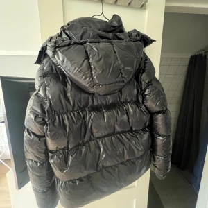 Svart/glansig pufferjacka - Svart pufferjacka från Everest med glansig yta och huva. 10/10 skick utan några defekter då den inte har använts. Den är storlek S men passar M också