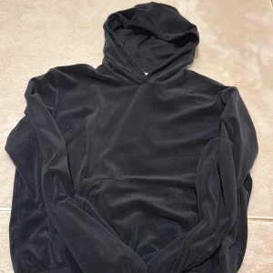 Svart hoodie i sammet från KappAhl - Mjuk och stilren svart hoodie från KappAhl i sammetsmaterial. Tröjan har huva, långa ärmar och en klassisk känguruficka framtill. Perfekt för dig som gillar en chill och snygg look.