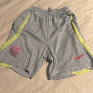 Grå PSG x Nike shorts med neon-detaljer - Säljer ett par grå PSG shorts från Nike med neon-gula detaljer längs sidorna och röd Nike-logga. Shortsens material är lätt polyester och de har elastisk midja med snörning. Perfekta för fotboll eller träning, riktigt snygga och sportiga.