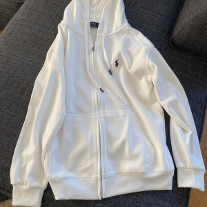  Polo Ralph Lauren Hoodie - Vit hoodie från Polo Ralph Lauren med dragkedja framtill och klassisk logga på bröstet. Tröjan har huva med snörning, ribbade muddar och fickor på sidorna. Perfekt för en chill och stilren look.