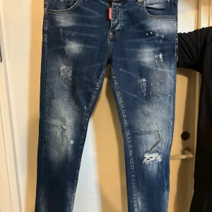 Dsquared2 blå distressed jeans - Snygga blå jeans från Dsquared2 med slitna detaljer, färgstänk och patchade hål för en riktigt edgy look. Klassisk femficksmodell med normal passform och tydlig Dsquared2-logga på röd etikett vid gylfen. Jeansen är tillverkade i denim med cool tvätt.