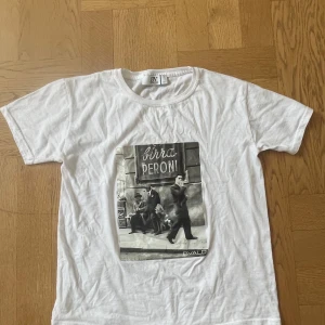 Vit t-shirt med Birra Peroni-tryck från Bvaldi - Vit t-shirt från Bvaldi i bomull med klassisk passform och korta ärmar. Framsidan har ett stort svartvitt fototryck med texten 'Birra Peroni' och motiv av personer på en gata. Perfekt för dig som gillar retrostil och coola prints.