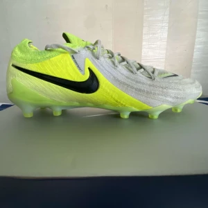Nike phantom GX ELITE fotbollsskor i gul och vit - Säljer ett par Nike Phantom GX ELITE  fotbollsskor i en fräsch kombination av neongult och vitt med svarta detaljer. Skorna har snörning, platt sula med dobbar av AG och är tillverkade i syntetmaterial för bästa grepp på planen. Det kommer även med en enkel väska. Perfekta för dig som vill ha så bra prestation som möjligt på fotbollen och som vill matcha med dina fotbollsskor och väskan.