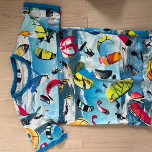 Färgglad t-shirt med kitesurfing från Molo - Cool t-shirt från Molo med färgstarkt kitesurfing-motiv över hela plagget. Basen är blå med inslag av gult, rött, grönt och svart. T-shirten har blå kantband och är kortärmad, perfekt för dig som gillar unika prints och sportiga vibes.