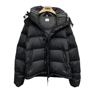 Svart pufferjacka från Burberry - Svart pufferjacka från Burberry med avtagbar huva och dragkedja framtill. Jackan har en klassisk pufferlook med breda kanaler, två fickor med dragkedja och gråmelerat foder med ränder. Perfekt för kalla dagar och riktigt snygg streetstil.