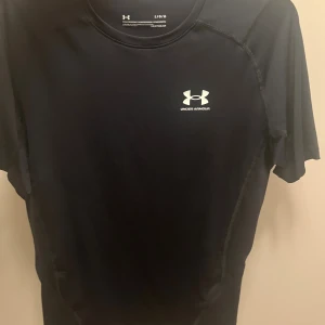 Svart Compresion shirt från Under Armour - Svart kortärmad Compresion shirt från Under Armour med logga på bröstet och ryggen. Tillverkad i ett stretchigt och lätt polyestermaterial som andas, perfekt för träning. Snygg passform och diskreta sömmar för en sportig look. 