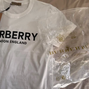 Vit Burberry t-shirt med logga - Vit t-shirt från Burberry med klassisk svart logga 'BURBERRY LONDON ENGLAND' tryckt framtill. T-shirten har rund halsringning och korta ärmar. Tillverkad i mjuk bomull och levereras med originalförpackning och tagg.