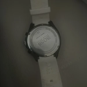 Svart och vit BOSS-klocka - Stilren klocka från BOSS med svart urtavla och vita detaljer. Klockan har en vit rem med BOSS-logga och flera små urtavlor för extra funktioner. Boetten är rund och i metall, medan remmen är i syntetmaterial. Perfekt accessoar för dig som gillar en clean och modern look. Priset kan diskuteras vid snabb affär