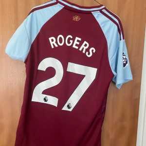 Aston Villa Rogers 27 - Oanvänd Aston Villa fotbollströja med Rogers 27 på ryggen. Slim fit-modell från Adidas i vinrött och ljusblått, med klubbmärke, Premier League-logga på ärmen och Betano som sponsor. Tillverkad i lätt och ventilerande polyester, perfekt för match eller träning.