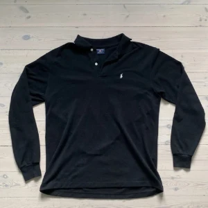 Svart långärmad pikétröja Polo Sport - Klassisk svart långärmad pikétröja från Polo Sport i 100% bomull. Tröjan har krage, två knappar framtill och den ikoniska broderade loggan på bröstet. Perfekt för en stilren och avslappnad look.