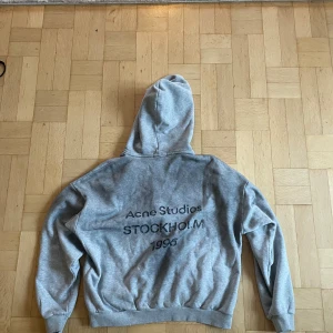 Grå hoodie från Acne Studios - Säljer en grå hoodie från Acne Studios med tryck på ryggen där det står 'Acne Studios STOCKHOLM 1996'. Tröjan har huva, ribbade muddar och är i mjuk bomull. Perfekt för en chill och stilren look. Ny pris 5300