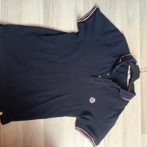 Mörkblå Moncler pikétröja - Snygg mörkblå pikétröja från Moncler med klassisk krage och korta ärmar. Tröjan har broderad Moncler-logga på bröstet och rödvita ränder på krage och ärmslut. Tillverkad i mjuk bomull som känns skön mot huden. Perfekt för en clean och sportig look.