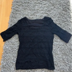 Svart spetsblus från Lindex - Svart blus från Lindex i storlek S med halvtransparent spets och rundad urringning. Toppen har trekvartsärm och är fodrad framtill och bak, men ärmarna är genombrutna. Perfekt för dig som gillar detaljerad spets och en klassisk look.