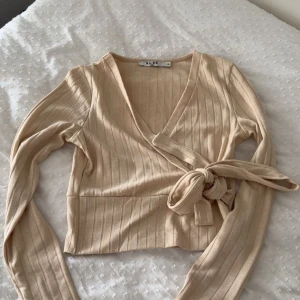 Beige omlottopp från NA-KD XS - Säljer en beige ribbad omlottopp från NA-KD i storlek XS. Toppen har långa ärmar, croppad passform och knyt i sidan. Den är tillverkad i mjuk bomullsmix och har en snygg v-ringning som ger en trendig look.